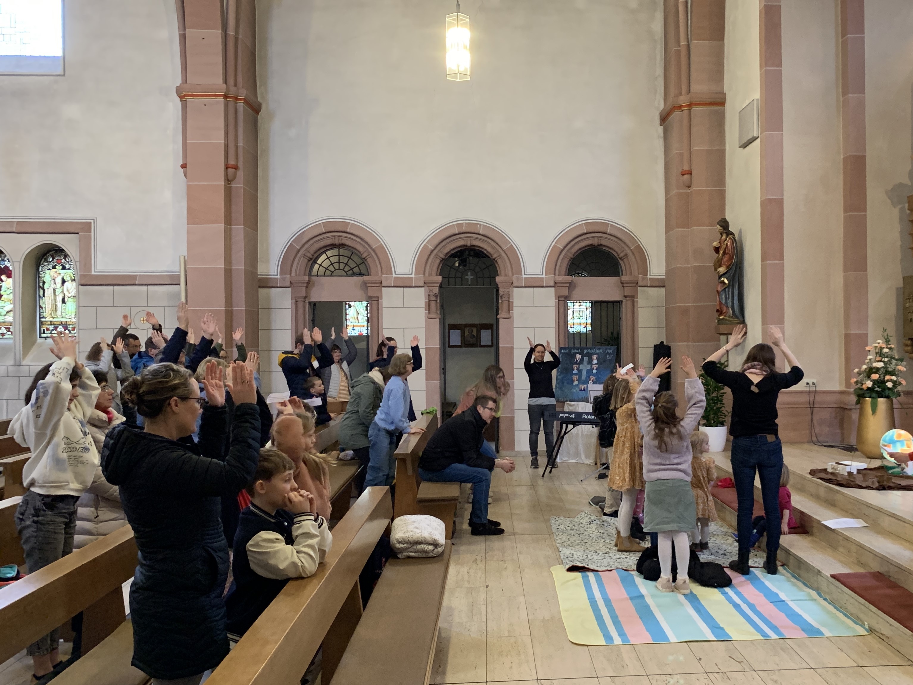 Menschen in Kirchenbänken heben die Arme. Kinder im Halbkreis auf dem Teppich ebenfalls.
