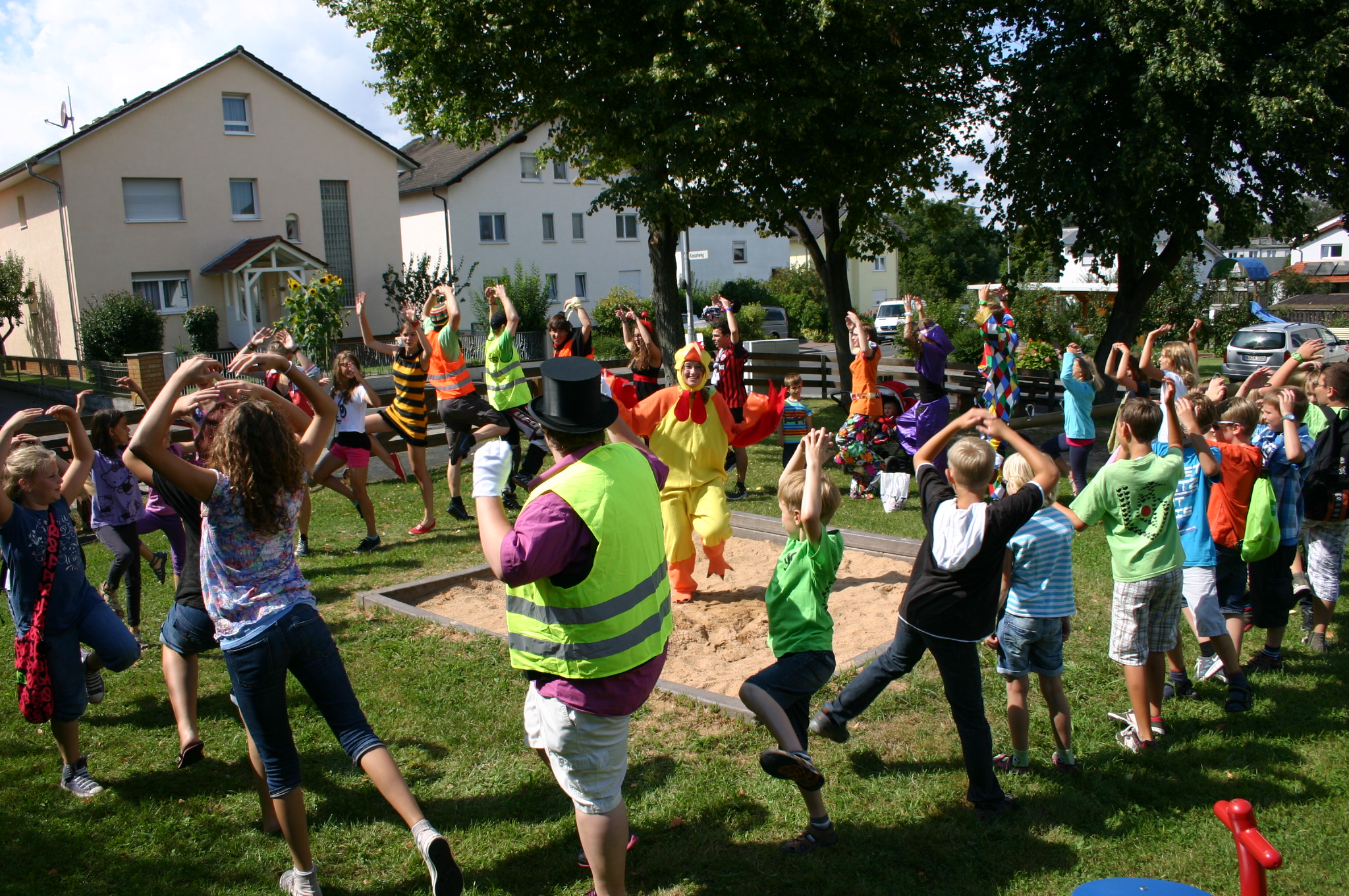 Viele Kinder und Jugendliche tanzen im Kreis. In der Mitte im Sandkasten tanz ein Jugendlicher im Entenkostüm. 