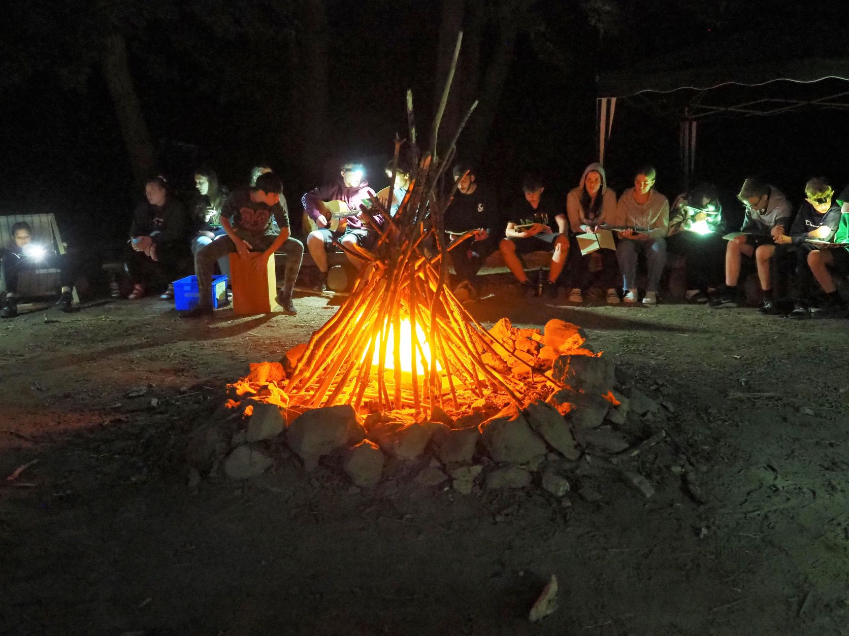 Lagerfeuer in der Mitte, im Hinterggrund sitzen Kinder und Jugendliche