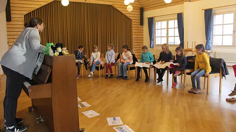 Chorleiterin am Klavier, Kinder sitzen um sie herum